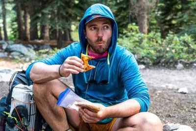 A person in a blue Ridge Merino Solstice Hoodie sits outdoors near a forest, holding a slice of pizza with a curious expression. They have a backpack beside them and a plastic bag in their lap, suggesting a relaxed, spontaneous meal in nature.