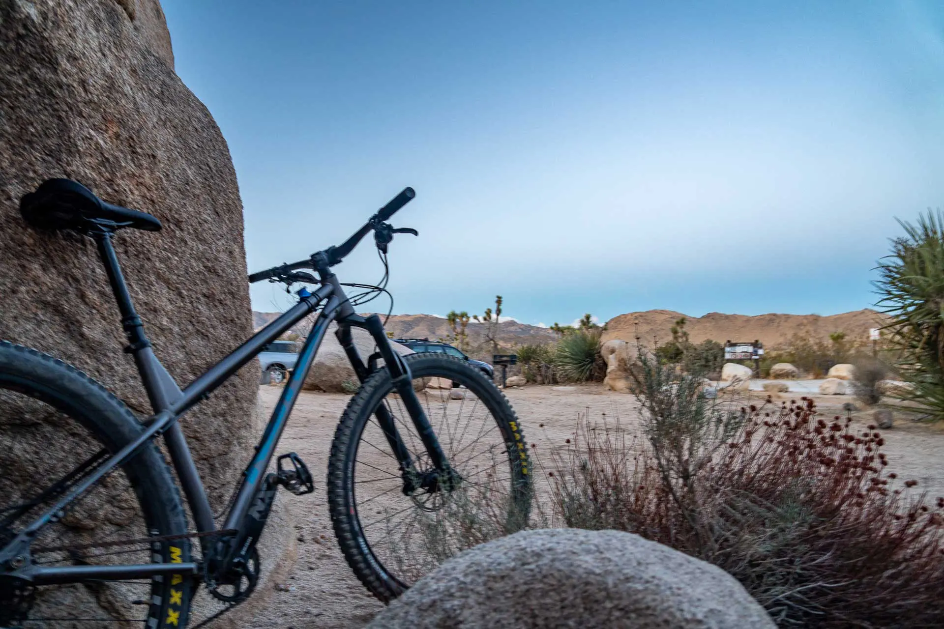A mountain bike, part of my essential AZT bikepacking gear list, leans against a large rock in the desert landscape. The sky is clear and blue, with distant hills and sparse vegetation including shrubs and small trees. Several parked cars dot the background.