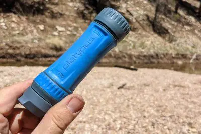 Close-up of a hand holding a Platypus Quickdraw water filter, its blue and gray cylindrical shape standing out against the river and rocky terrain in the background.