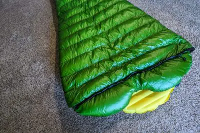 A green sleeping bag is laid out on a beige carpet. Inside, a yellow inflatable sleeping pad is visible, revealing the open end of the Western Mountaineering Astralite sleeping bag. The scene suggests a setup for indoor camping or testing camping gear.