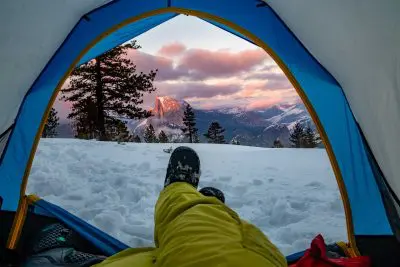From inside a Sierra Designs Convert 2 tent in snowy mountains, a person in yellow pants and boots is lying comfortably. The tent perfectly frames a picturesque scene of pine trees and a mountain peak under the colorful, cloudy sunset sky.