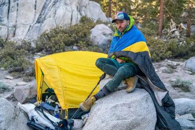 A person sits on a rock in a campsite, wrapped in a blue and yellow flag. Behind them is a Sierra Designs Nitro Quilt near the yellow tent, surrounded by trees and rocks. They wear a cap and casual outdoor clothing, embracing the serene woodland setting.