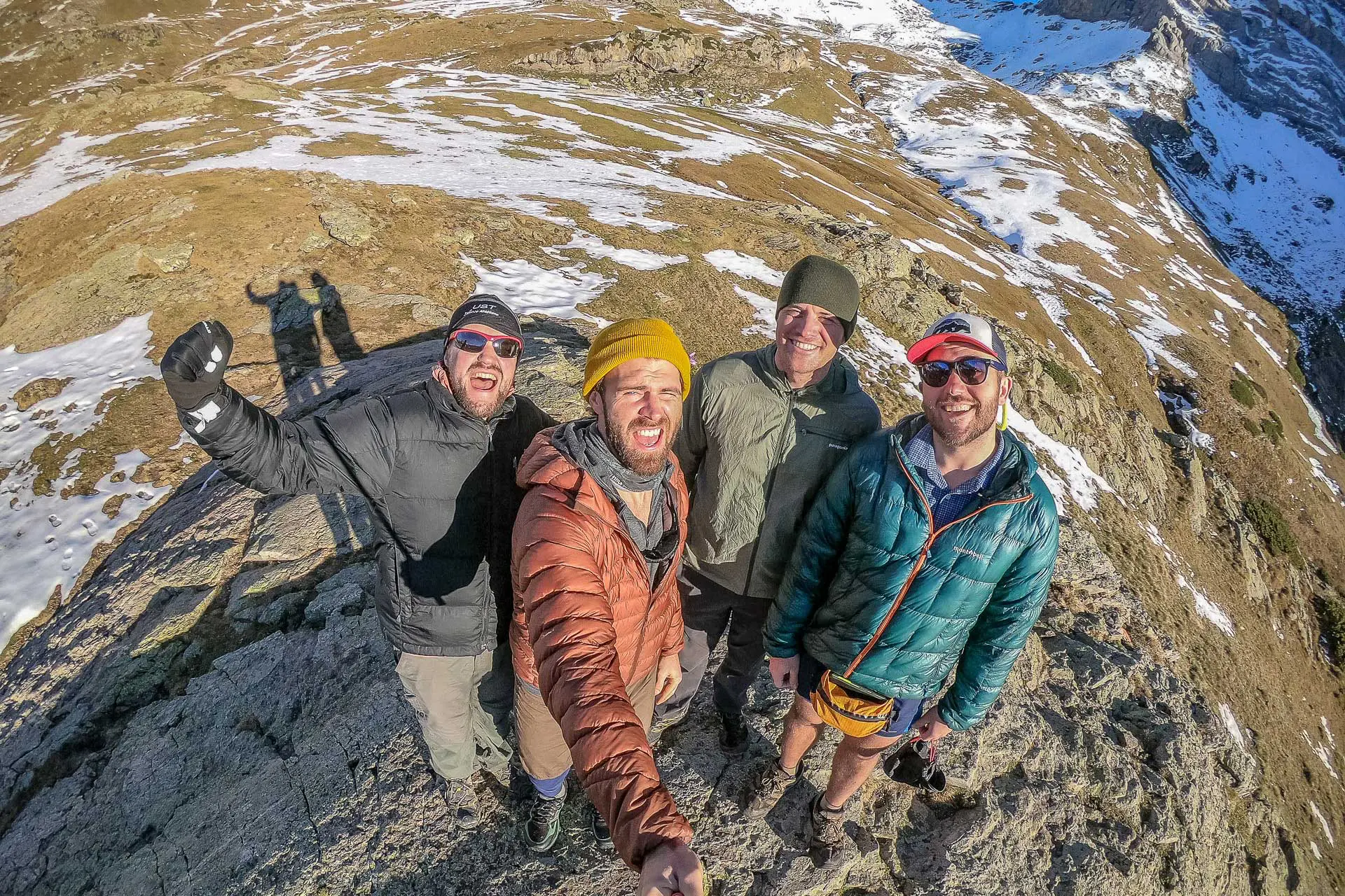 Four men, part of a tramily, smile and pose on a rocky mountain peak. They wear jackets and hats, casting shadows on the ground. In the background, snowy patches and rugged terrain stretch beneath a clear blue sky.
