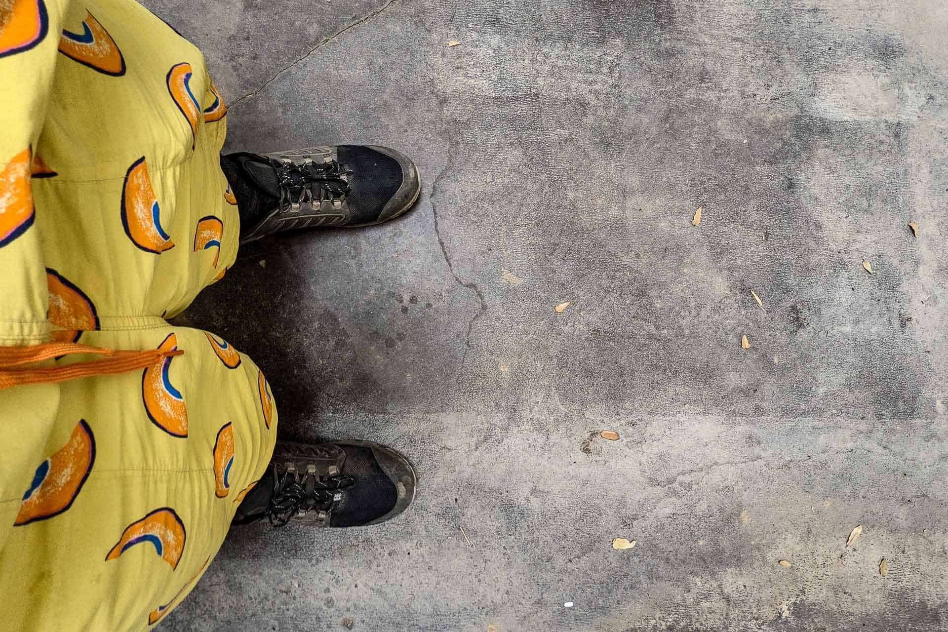 A person wearing black sneakers and yellow Patagonia Baggies with an orange pattern stands on a textured, gray concrete floor scattered with small leaves. The image is taken from an overhead perspective.