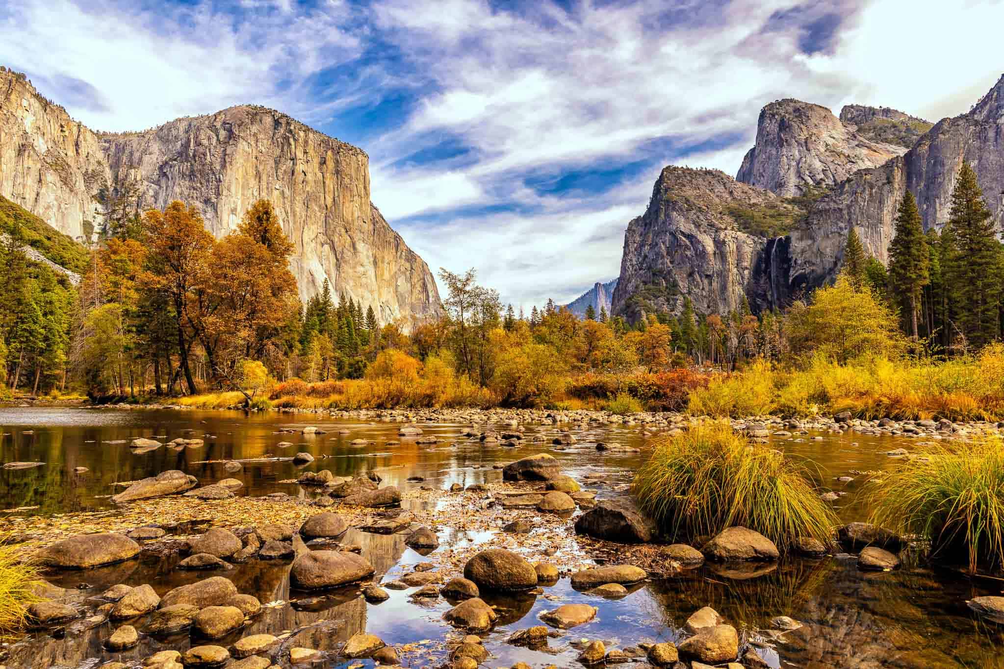 A scenic view of Yosemite Valley with rocky cliffs and autumn foliage reflecting in a calm river, captured during a John Muir Trail survey. The sky is partly cloudy, and evergreen trees line the banks, adding contrast to the vibrant fall colors.