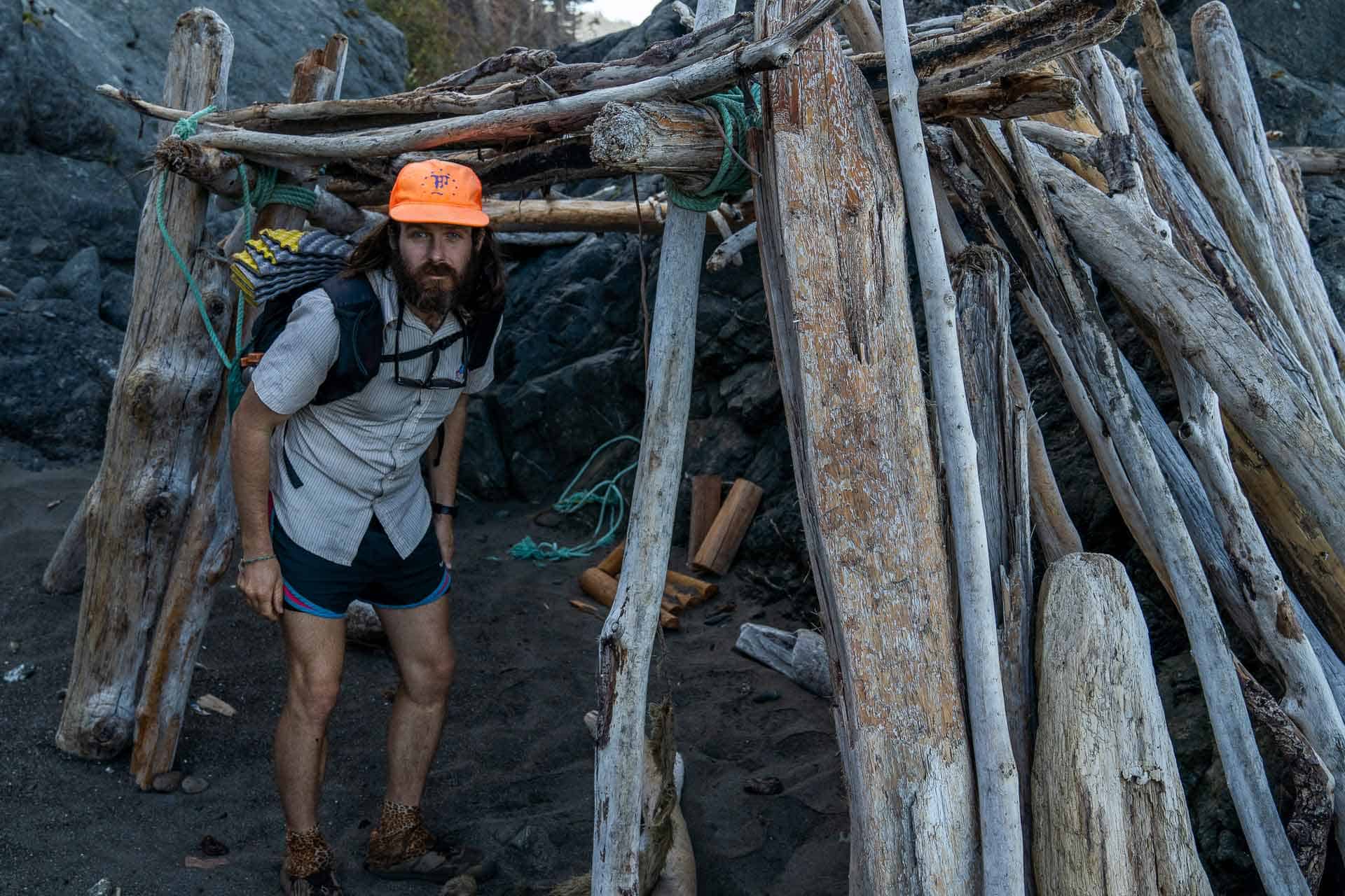 A man wearing a bright orange cap and carrying a backpack crouches under a driftwood shelter on a rocky beach, contemplating the importance of self-reliance and learning to stop depending on trail angels. He is dressed in a light shirt and shorts, surrounded by large rocks and pieces of driftwood.