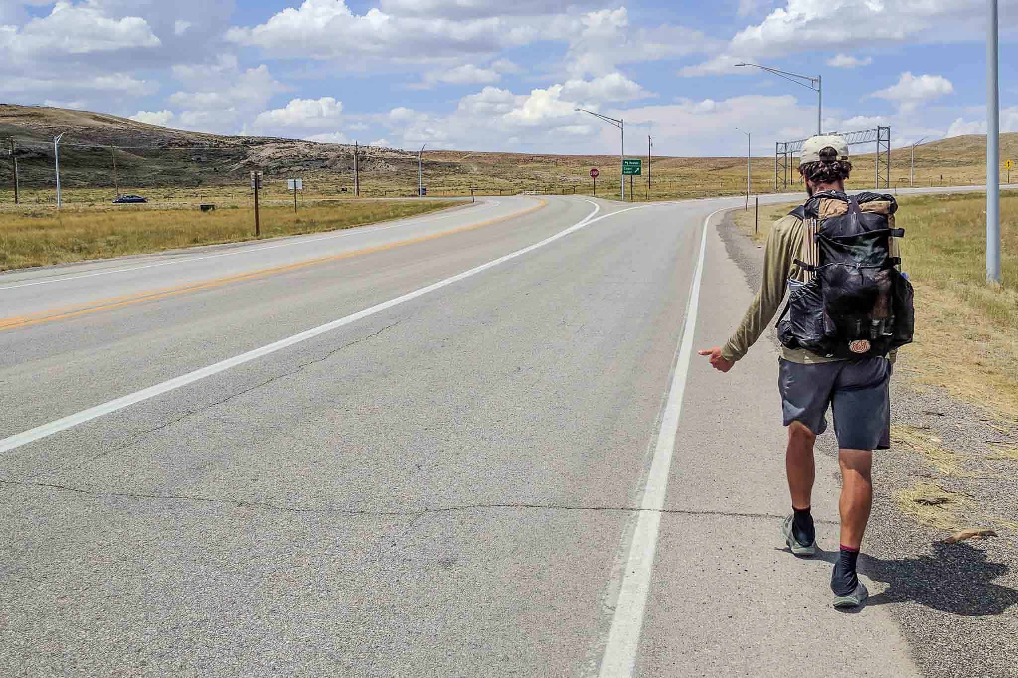 A person with a backpack walks along the side of a highway under a partly cloudy sky, much like a solitary hiker on the CDT. The road curves ahead, signs visible in the distance. The open, grassy landscape stretches endlessly as they gesture with their right hand.