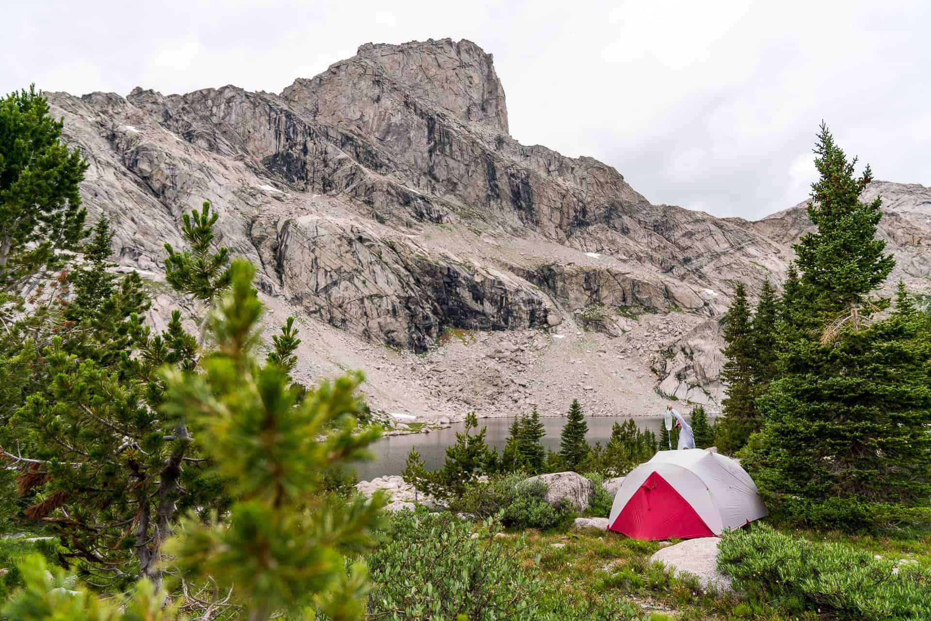 A red and white tent, resembling CDT survey gear, is set up near a small lake with a rocky mountain in the background. The foreground is filled with green trees and shrubs under a cloudy sky.