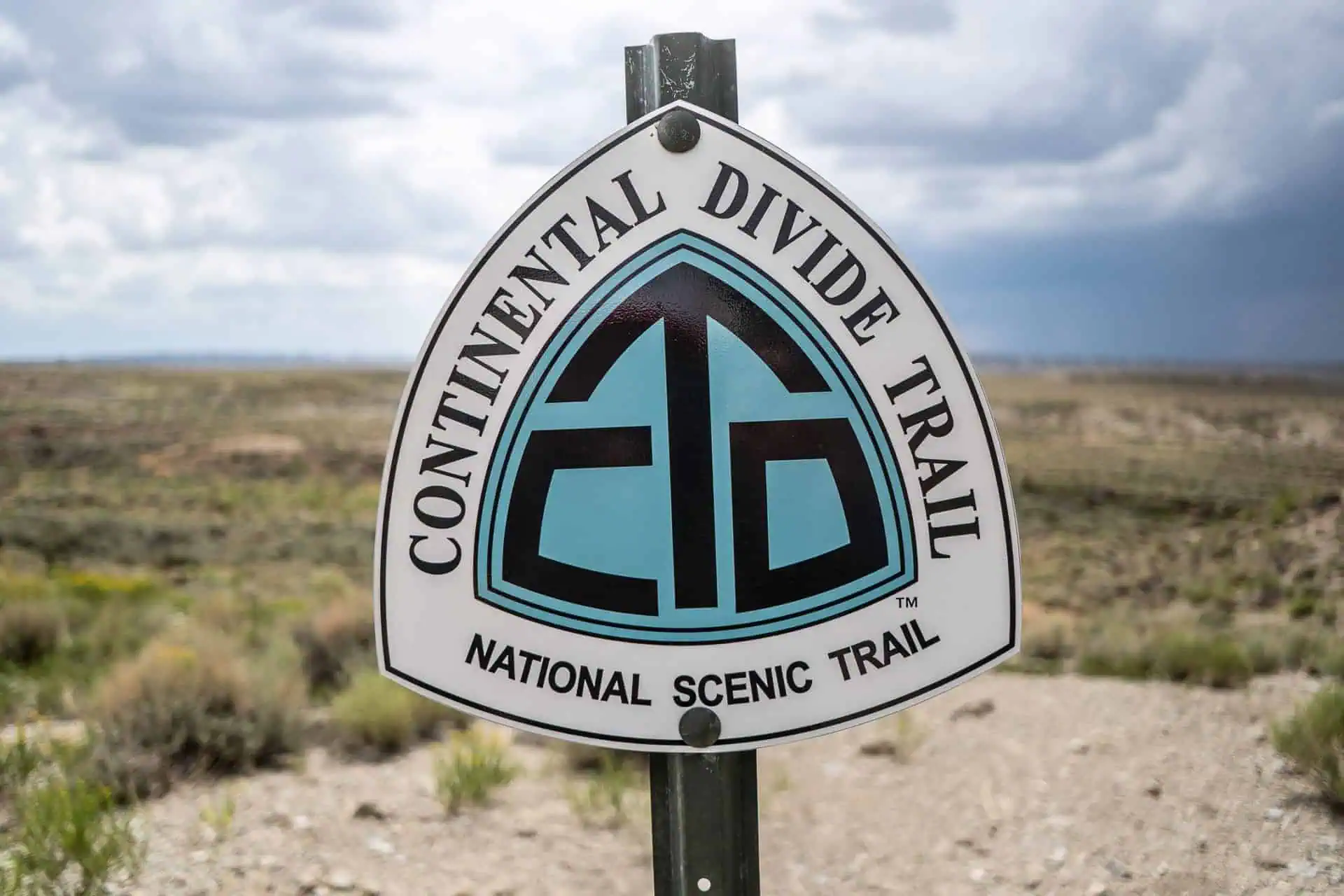 A sign for the Continental Divide Trail, documented in the CDT Survey 2021, stands against a vast, cloudy landscape. It proudly displays the trails logo in blue and black. The sky is overcast, complementing the open and rugged terrain of this National Scenic Trail.