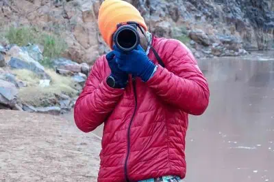 A person in a red jacket, possibly a Patagonia Micro Puff, and an orange beanie is snapping a photo with a large camera lens. They stand by a rocky riverbank with brown water coursing past. Blue gloves and a smartwatch complete their adventurous ensemble.