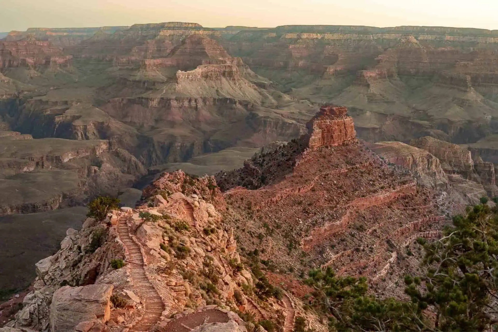 A scenic view of the Grand Canyon at sunset, showcasing a rugged landscape with layered rock formations and deep valleys. The sun casts a warm glow on the reddish-brown cliffs, while the famous R2R2R trail winds through this breathtaking terrain.