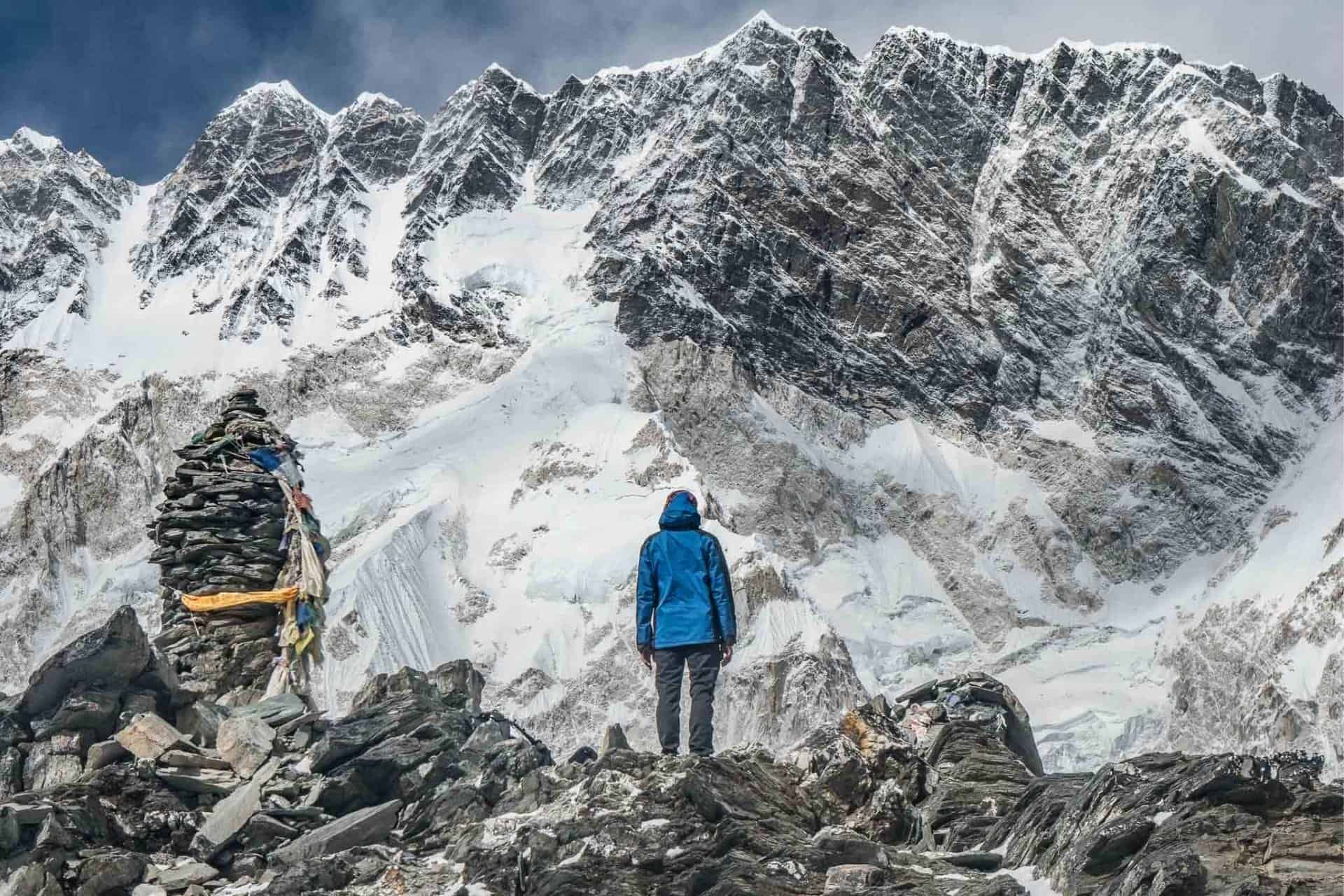 A person in a blue jacket stands on rocky terrain, facing the towering, snow-covered peaks of the Great Himalaya Trail. A small cairn of rocks with tied cloth is nearby, beneath a cloudy sky.