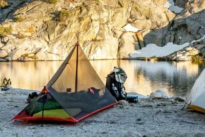 A person is resting inside a triangular mesh SlingFin SplitWing tent on rocky ground beside a tranquil lake. A backpack and supplies are outside the tent. Sunlit cliffs and patches of snow surround the water, reflecting the serene outdoor setting.