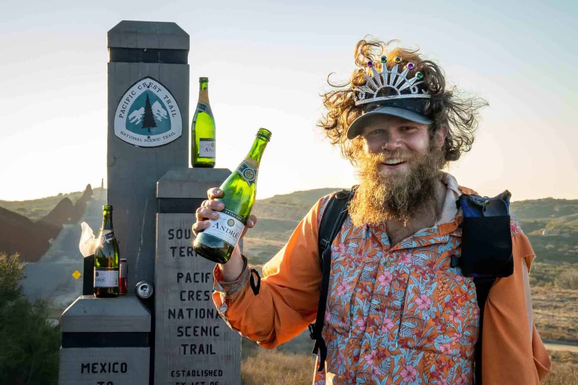 A person with a beard, crowned and donned in bright orange attire, holds a bottle of sparkling wine. Standing at the southern terminus monument of the Pacific Crest Trail, used in the 2022 PCT hiker survey, they are framed by scenic hills in the background.