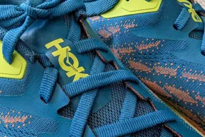 Close-up of a Hoka One One Speedgoat 5 running shoe in blue and orange with teal laces. The shoe boasts a bold yellow brand logo and textured mesh fabric, enhanced by green accents near the laces for added contrast.