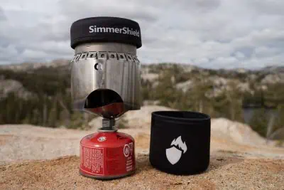 A portable camping stove with an innovative SimmerShield and a red fuel canister rests on a rocky surface. Beside it lies a black protective sleeve adorned with a flame logo. The backdrop showcases a blurred, rocky landscape dotted with sparse trees under a cloudy sky.