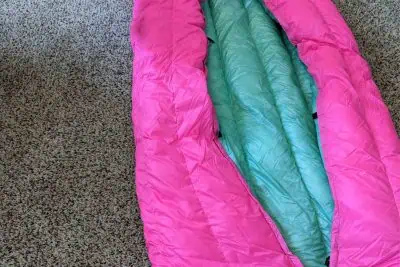 A pink and mint green sleeping bag, resembling the colors of the vibrant ugq bandit gear, is partially unzipped on a textured carpet. The outer layer is pink, and the inner lining is mint green. The beige carpet beneath features a speckled pattern.