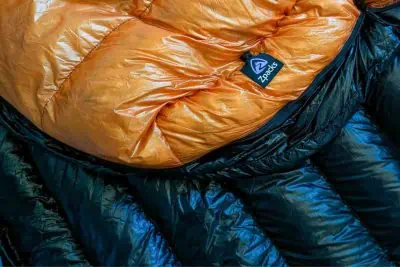 Close-up of a shiny, crumpled orange and black Zpacks sleeping bag with a visible brand label. The texture appears smooth and glossy, hinting at a weather-resistant material thats perfect for zip-around comfort in the great outdoors.