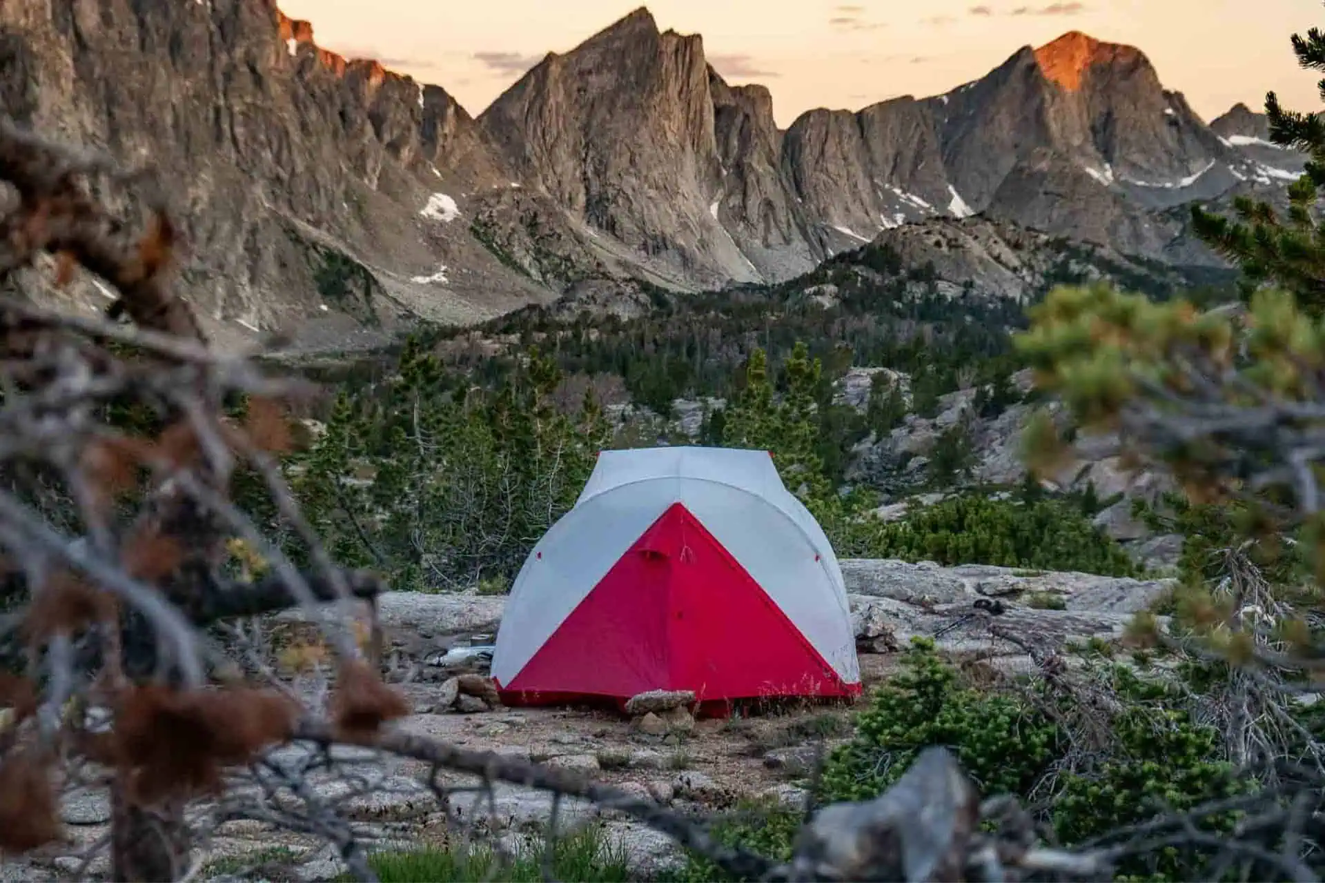 A red and white tent from the CDT Gear Guide 2022 is set up on rocky terrain, surrounded by pine trees. In the background, a range of rugged, sunlit mountains with patches of snow rises against a clear sky at sunset.