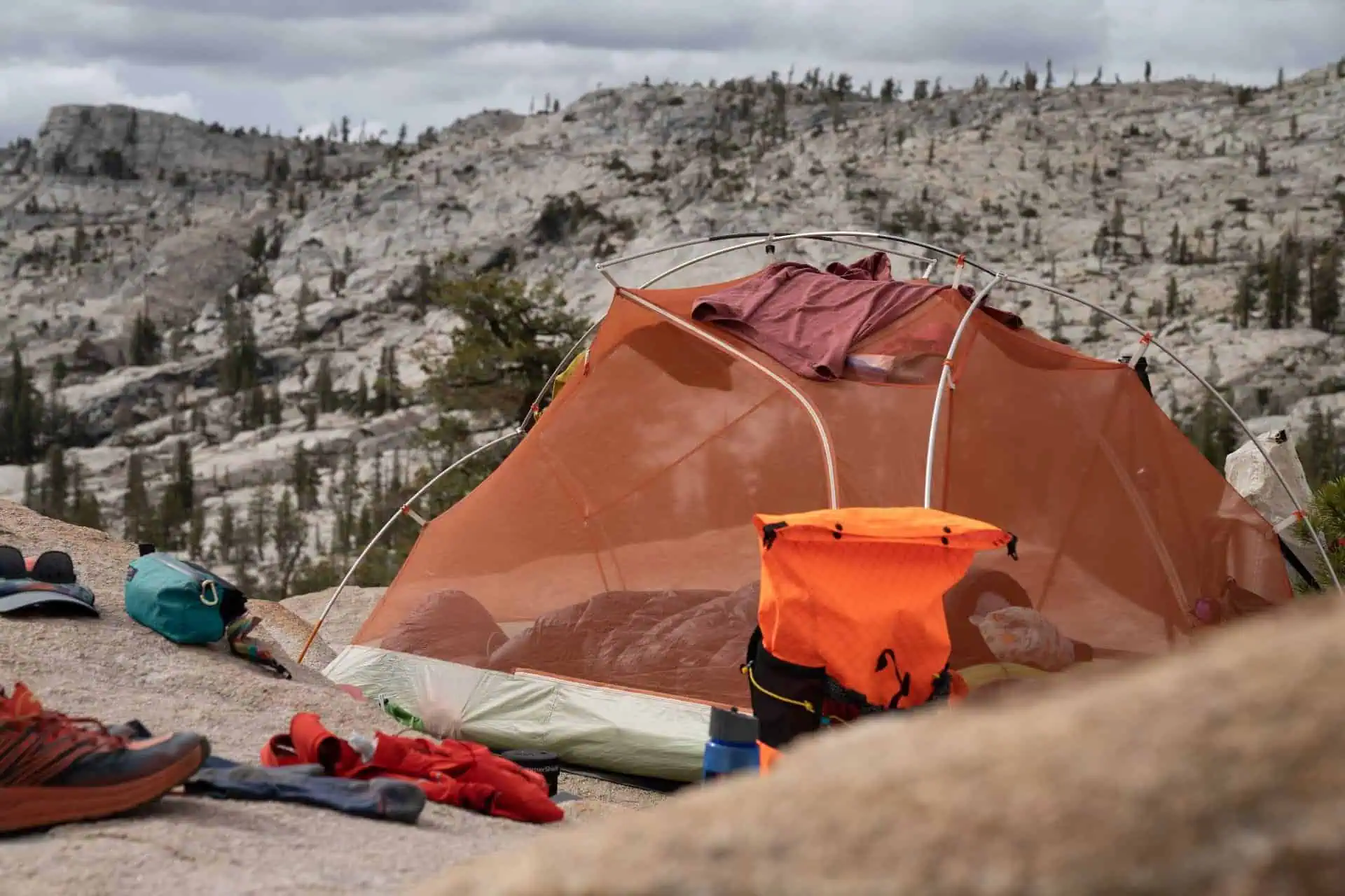 A camping scene unfolds on rocky terrain with a transparent tent. Clothes drape over it as if having just perused a PCT gear guide, 2022 edition. Scattered items, including an orange backpack and shoes, blend seamlessly into the landscape of pine trees and rocky hills beneath a cloudy sky.