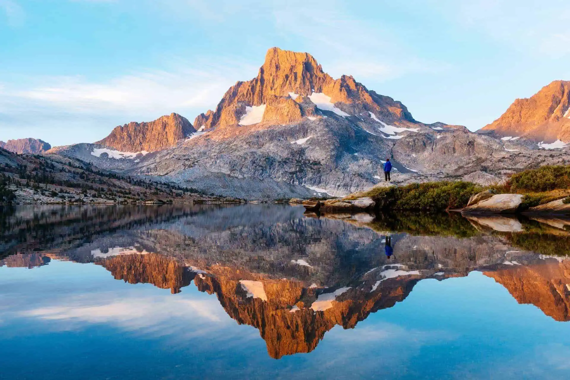 A person stands on a rock by a calm lake, equipped with the latest JMT Gear Guide 2023 essentials. A towering mountain, its snowy patches reflecting in the water, basks in warm sunlight under a clear blue sky.