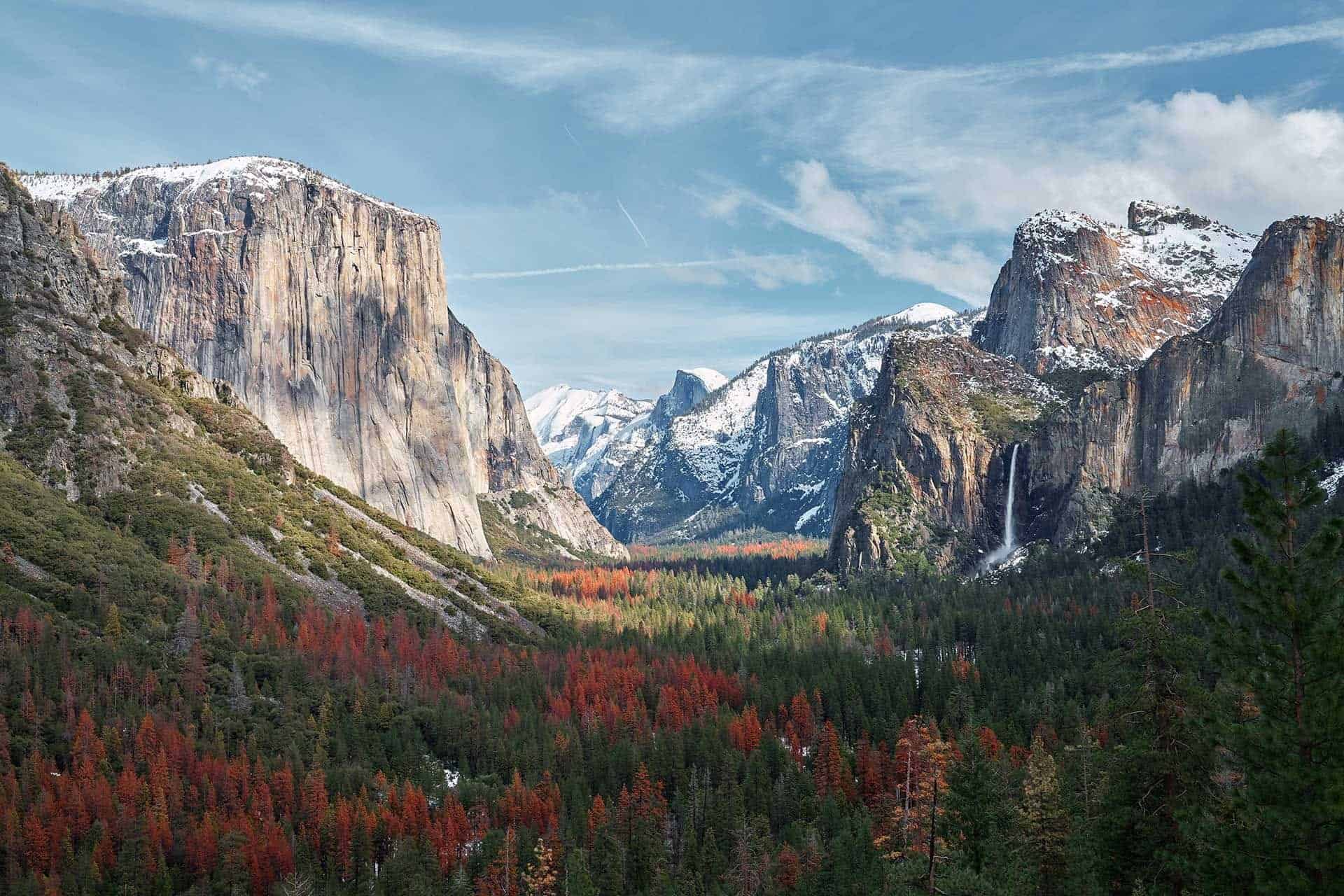A scenic view of Yosemite Valley showcases the awe-inspiring beauty captured during the 2022 John Muir Trail survey, featuring towering cliffs, a winding river, snow-capped mountains, and forests with autumn-colored trees. A waterfall cascades down one side under a partly cloudy sky.