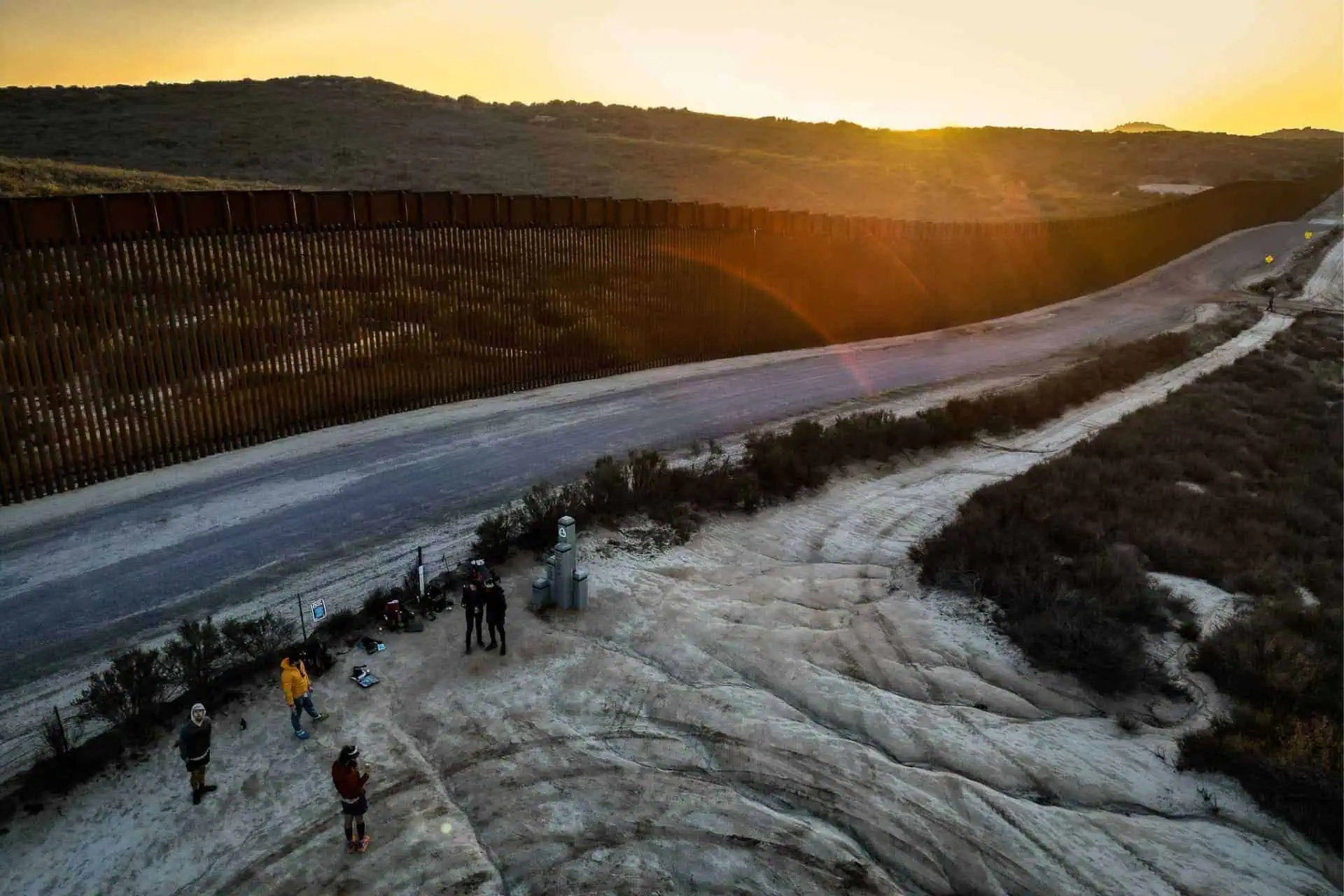 Aerial view of a border landscape at sunset with a long fence stretching across the image. Near equipment on the sandy, rocky area in the foreground, several people gather, possibly exchanging Pacific Crest Trail hiking advice as the golden sky casts a warm light over the scene.