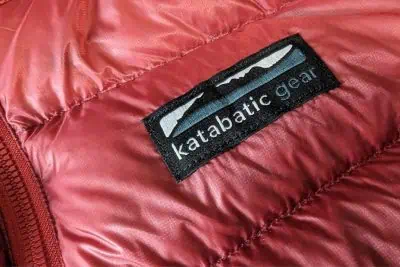 Close-up of the Katabatic Tincup Down Jacket, a red puffer featuring a black label with katabatic gear text and a stylized mountain graphic. The visible stitching and fabric texture add intricate detail to the garments design.