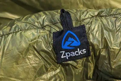 Close-up of a green fabric featuring a black tag with the blue and white Zpacks logo, showing a stylized mountain design. The tag, stitched into the fabric of a Zpacks mummy sleeping bag, reveals the fine texture and durable construction.