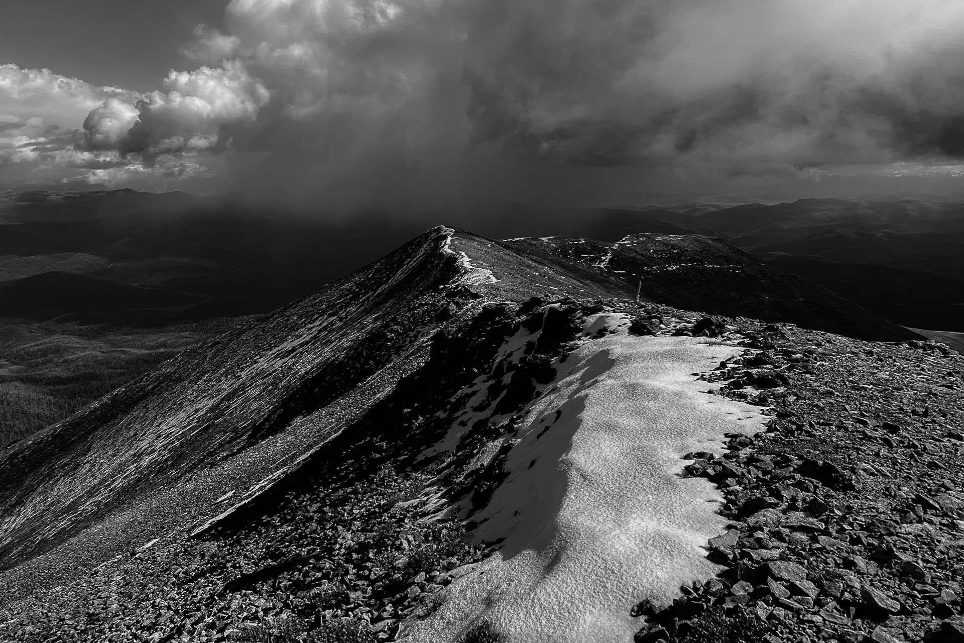 The black-and-white image of a rugged mountain ridge, reminiscent of CDTs horror stories, displays a snow-covered path leading into a dark, moody sky with dramatic cloud formations.
