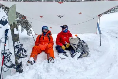 Two people sit under a white tarp in the snow, dressed in Arcteryx Rush Bib Pants and bright snow gear. Snowboards and a backpack lie nearby. Relaxed amidst snow-covered trees, theyve created a cozy winter campsite.