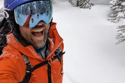 A person in an Arcteryx Beta jacket and blue helmet is excitedly skiing through a snowy landscape. Their mirrored goggles reflect the scenery, enhancing the experience. Snow-covered trees are visible in the background, adding to the serene winter tableau.