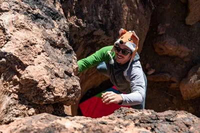 A person in a skygoat camp hoodie and sunglasses climbs between rocks in a rugged outdoor setting, looking up with a smile. The sunny scene highlights the earthy tones of the rocks and their casual attire, topped with a playful animal hat.