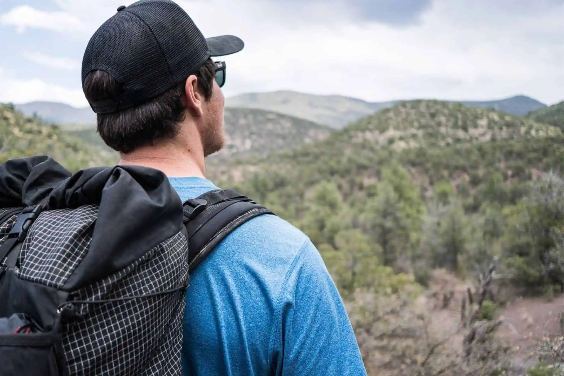 A person wearing a cap, sunglasses, and a blue shirt stands outdoors, backpack slung over their shoulder. They gaze at the landscape of rolling hills and greenery under a cloudy sky, contemplating their next adventure on one of the most popular trail names in the area.
