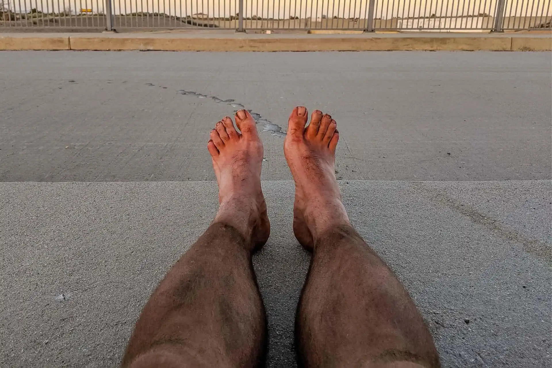 Dirty feet and legs of a person lying on a concrete surface, with a metal railing and sky in the background, likely taking a breather after conquering a trail. The person appears to be resting or relaxing outdoors.