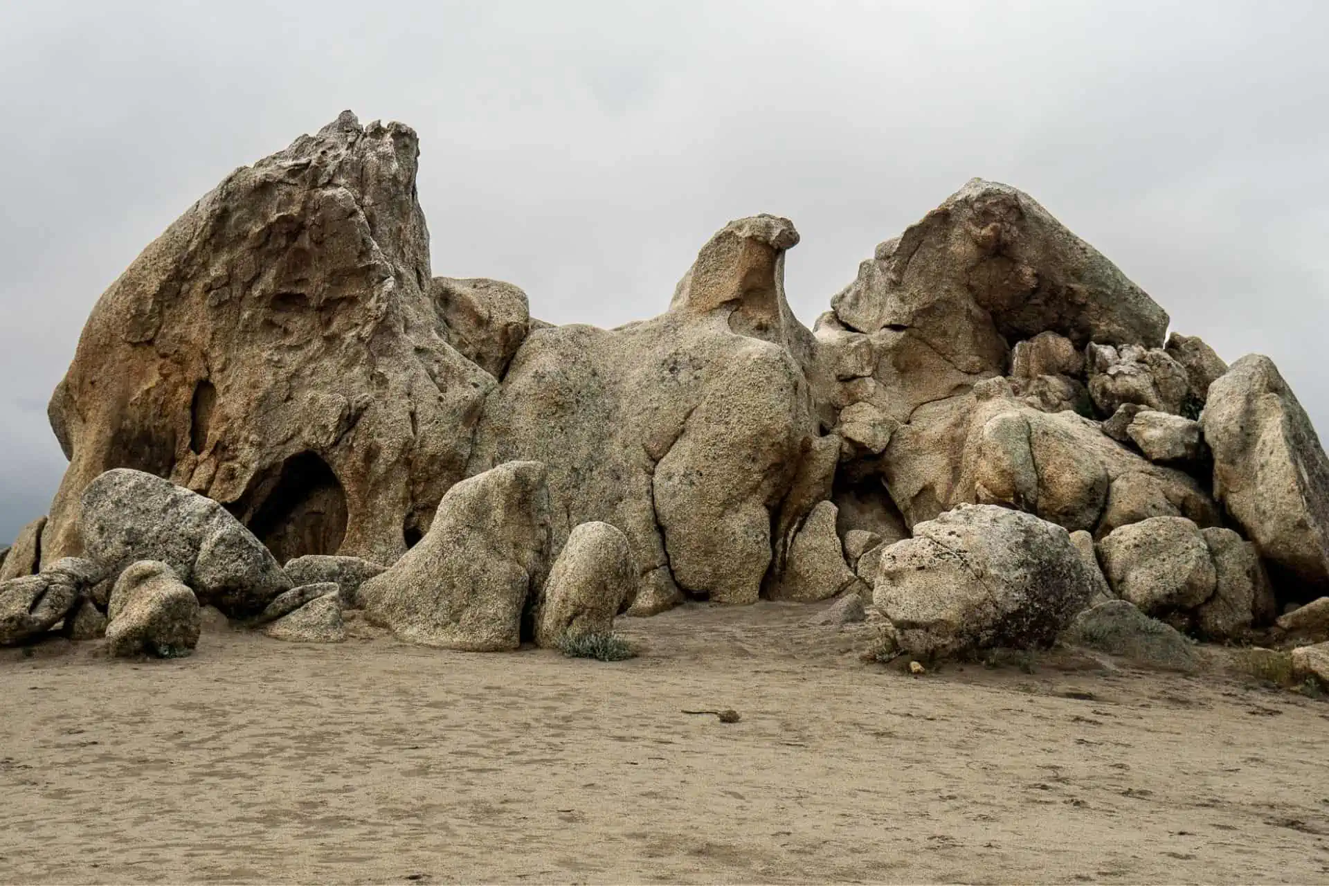 A rugged formation of large, weathered boulders and rocks on a sandy surface, reminiscent of the best PCT moments 2022, set against a cloudy sky. The rocks have varied textures and shapes, suggesting erosion over time.