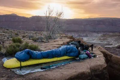 A person in a blue sleeping bag rests on a Therm-a-Rest Xlite NXT atop a rocky ledge at sunset. They are surrounded by camping gear, with a tree and distant cliffs visible under the glowing sky.