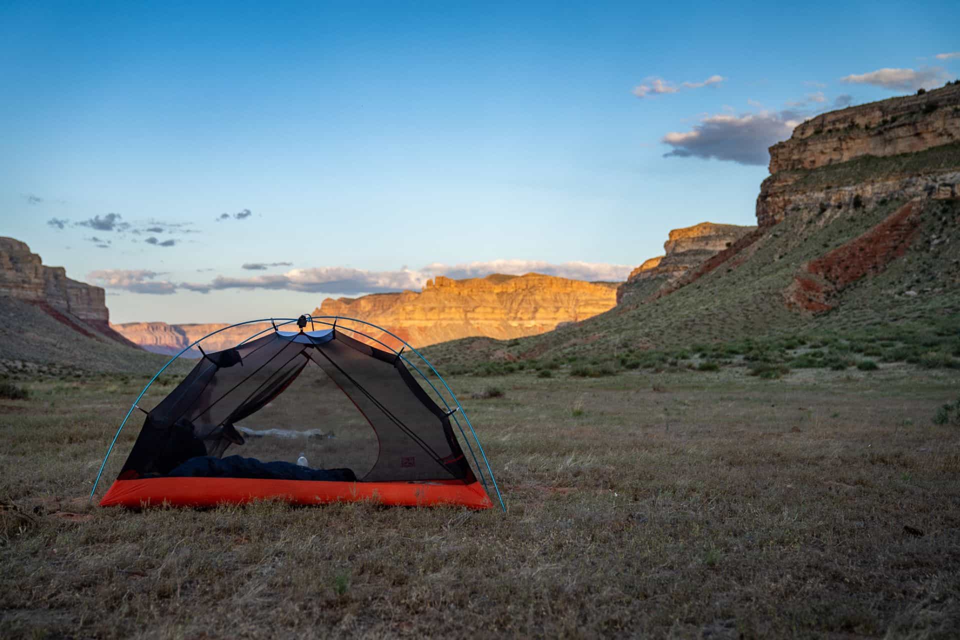 A SlingFin Portal 1 tent, in vibrant orange and gray, is set up on a grassy plain surrounded by rugged cliffs and hills. The sun casts a warm glow on the rocks, while the mostly clear sky is dotted with a few clouds.