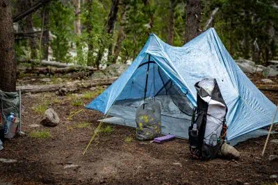 A small Zpacks Duplex Zip Tent is set up in a forested area, accompanied by a backpack and camping gear. The ground is strewn with leaves and small rocks, while trees and lush greenery form a serene backdrop.
