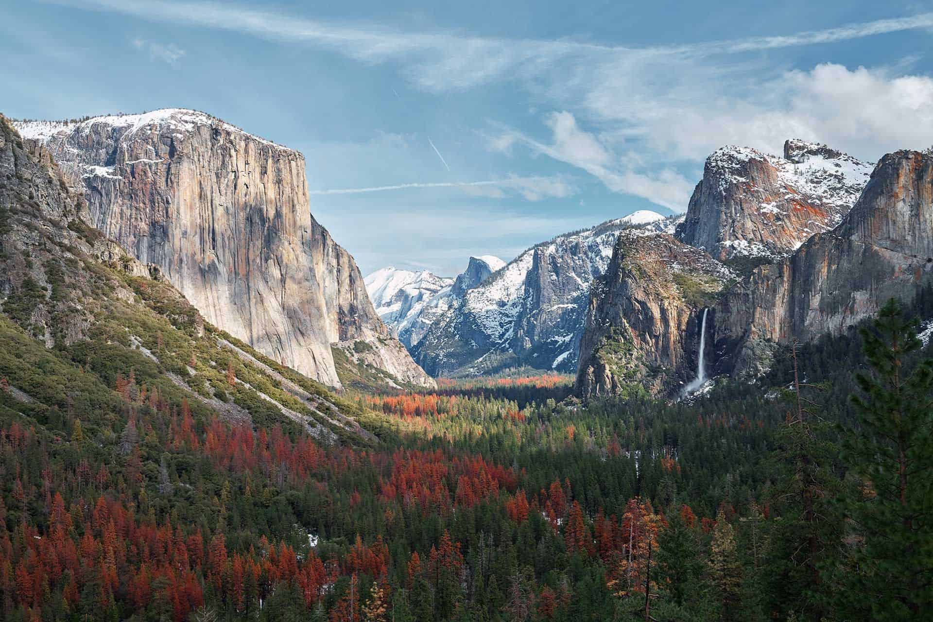 A breathtaking view of Yosemite Valley showcasing towering granite cliffs and snow-capped peaks under a blue sky. A dense forest with trees in autumn hues stretches in the valley with a waterfall visible on the right.