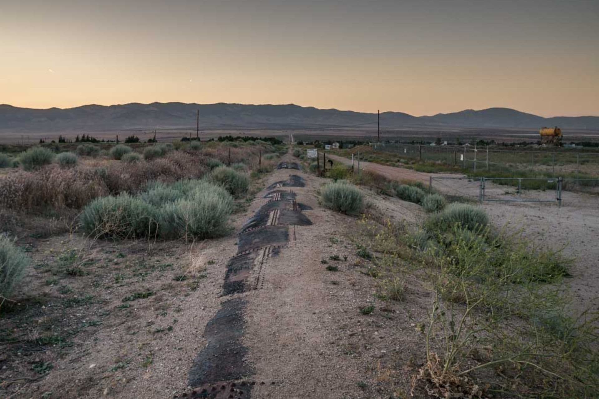 A deserted rural landscape at sunset reveals a worn road leading into the distance, reminiscent of a forgotten side quest on the Pacific Crest Trail. The path is fringed by dry grass and sparse vegetation, with mountains looming on the horizon under a dimming sky.