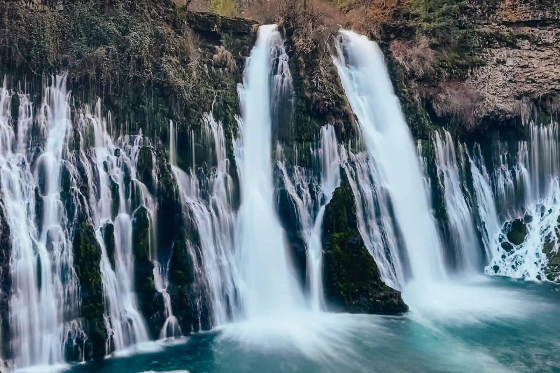 A scenic waterfall cascades over a rock face adorned with green moss and vegetation, mist rising from multiple streams above the blue pool below. This lush, natural wonder is one of the Pacific Crest Trail side quests in Northern California.