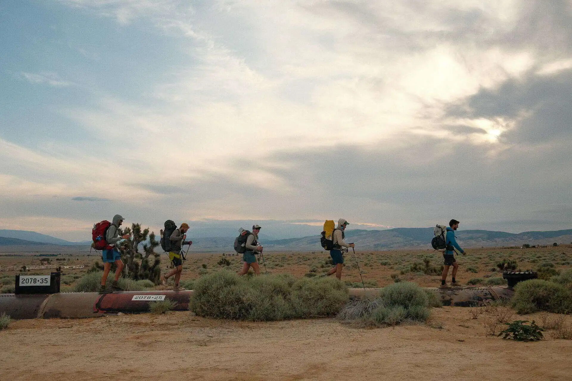 A group of people, equipped with the latest PCT gear, hiking on pipes across a vast desert landscape. Theyre carrying backpacks and using poles, with the horizon and a cloudy sky in the background. The terrain is sandy with some shrubs scattered around.