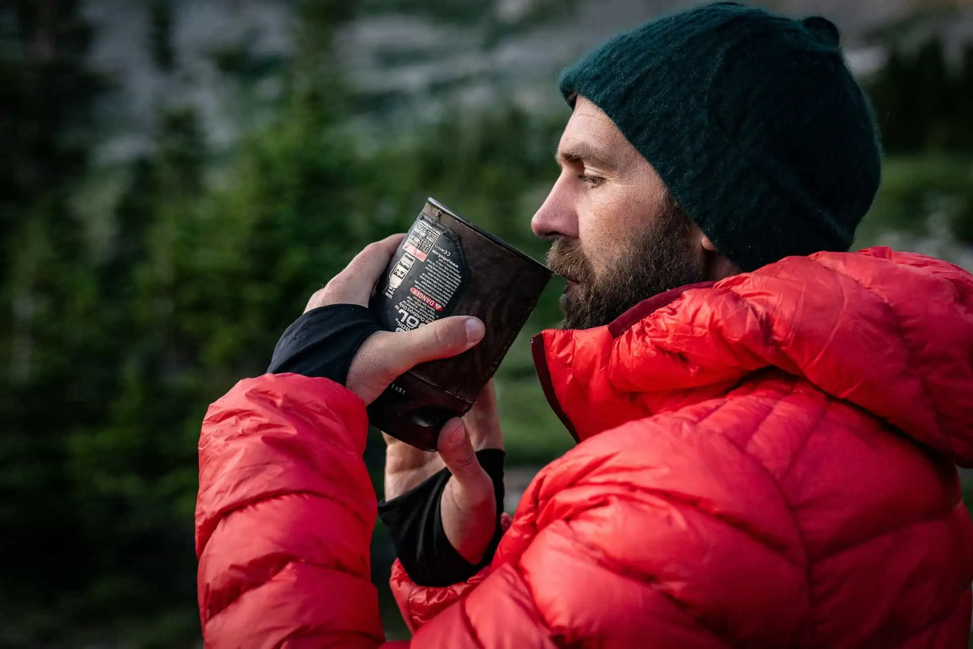 Amidst the natural backdrop of blurred greenery, a person in a red puffer jacket and dark beanie sips from a cup outdoors. The scene evokes the serene spirit found in guides like the CDT Resupply Guide 2023, perfectly capturing an adventure-ready moment.