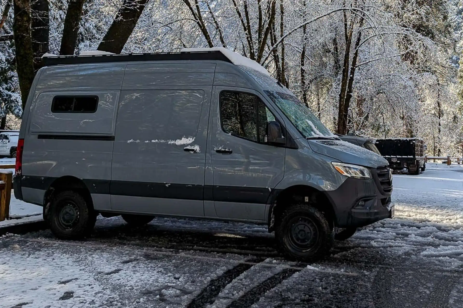 A grey van is parked on a snowy road, alongside snow-covered trees, forming the first step in this serene wintery tableau. The sky is clear, enhancing the crisp beauty of this tranquil setting.