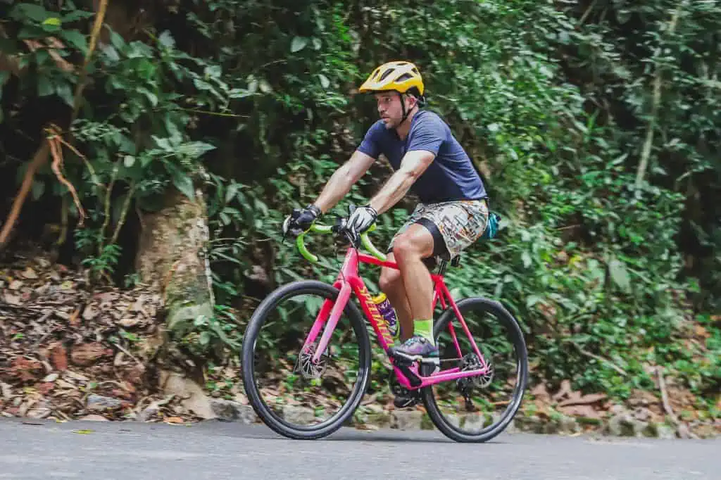 A guy in a yellow helmet riding a pink bicycle up a hill