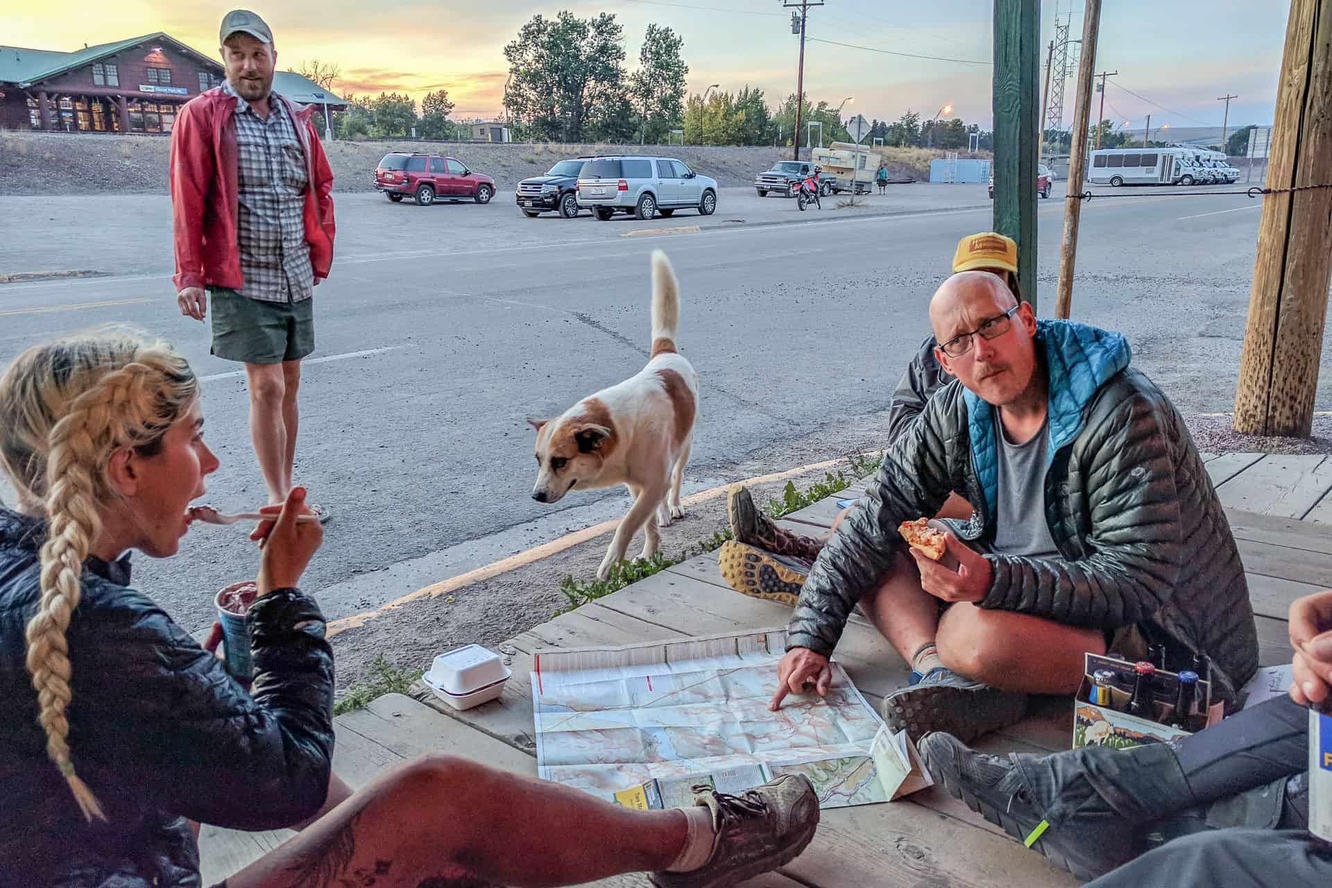 As the sun sets over this rural scene, marking one of the cdt best moments of 2023, a group casually gathers on a porch. They are immersed in reading a map while a dog strolls by. In the background, someone stands watchfully as the cars line up across the street.