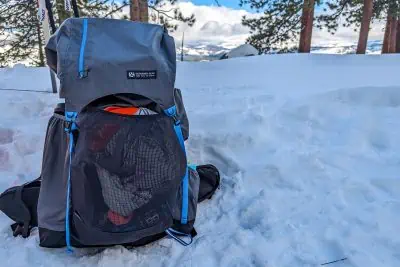 A gray and blue Gossamer Gear Mariposa backpack sits in the snow against a backdrop of pine trees and a mountainous landscape under a partly cloudy sky, evoking a sense of adventure and freedom.