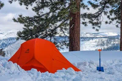 An orange MSR Access 2 tent is pitched on a snow-covered landscape surrounded by tall pine trees. A blue snow shovel is stuck in the ground nearby. In the background, snow-capped mountains stretch under a partly cloudy sky, showcasing a perfect winter camping experience.