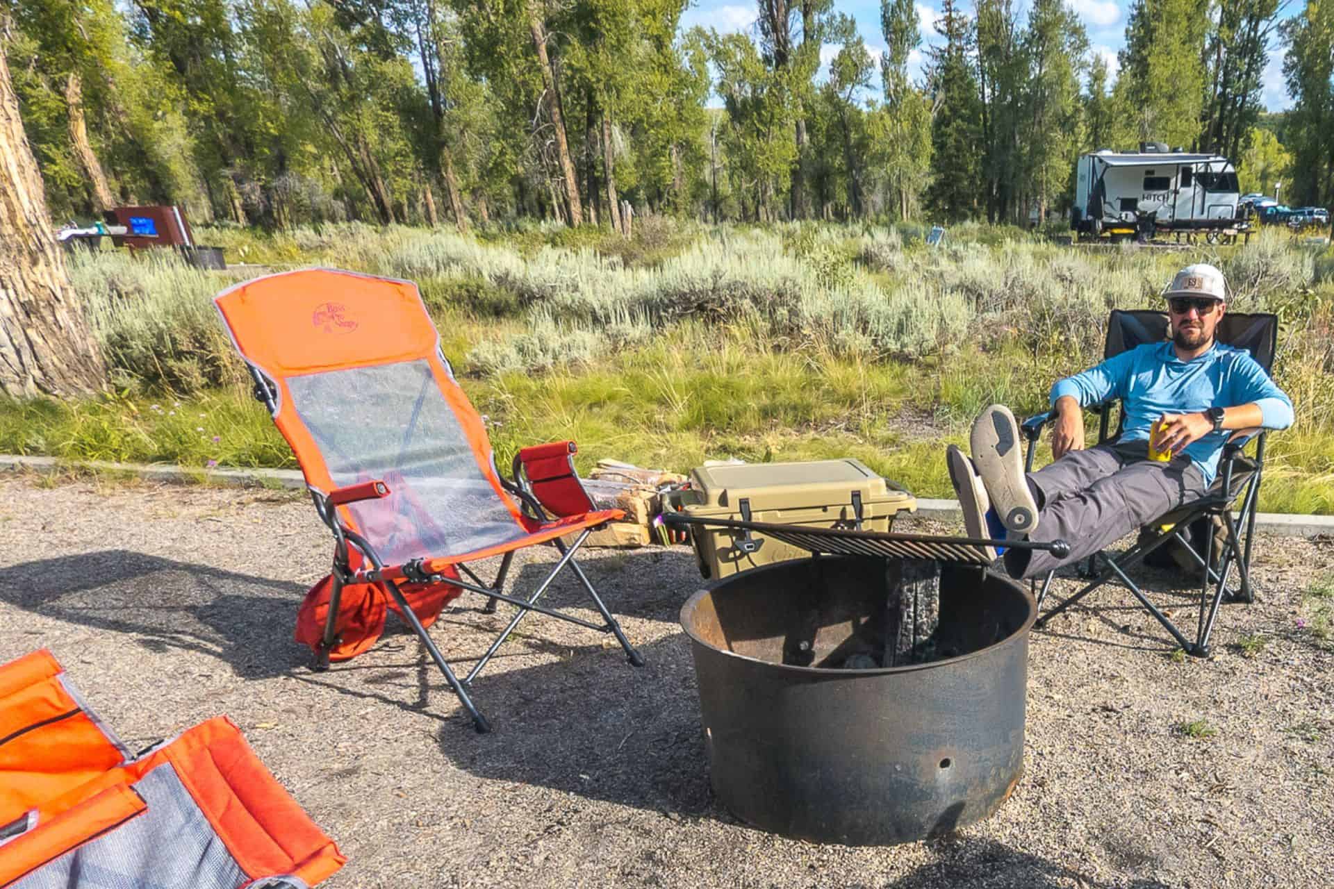 A man wearing sunglasses and a hat relaxes in a camping chair with his feet on a fire pit at a scenic campground. Nearby, an orange chair waits as if trail angels have left it behind. A camper and trees fill the backdrop under the expansive blue sky.
